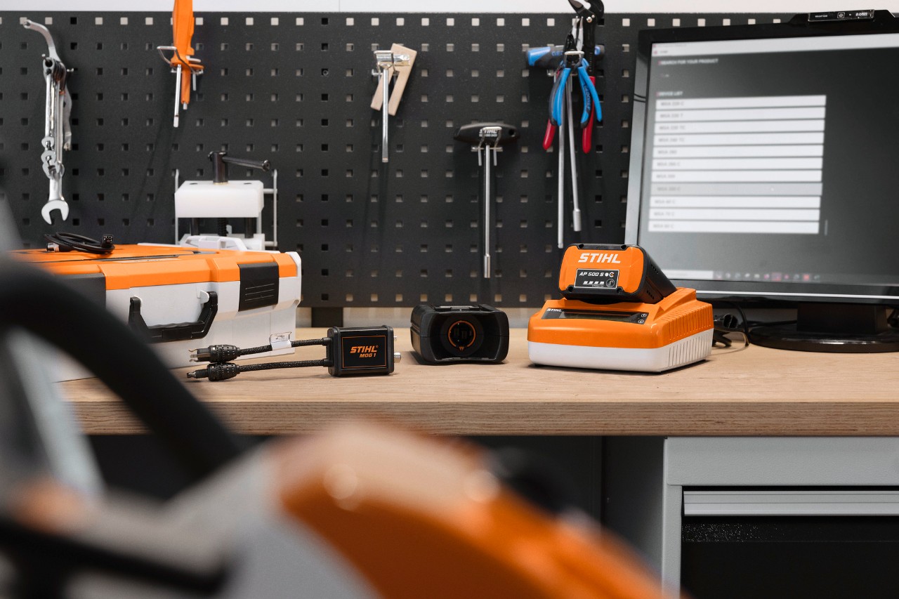 In a workshop, a man stands in the foreground operating a PC. On the table in front of him is a chainsaw