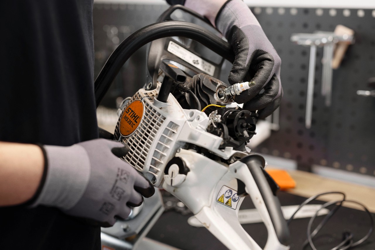 In a workshop, a person wearing gloves is standing in the foreground, holding the engine of a chainsaw and changing a spark plug.