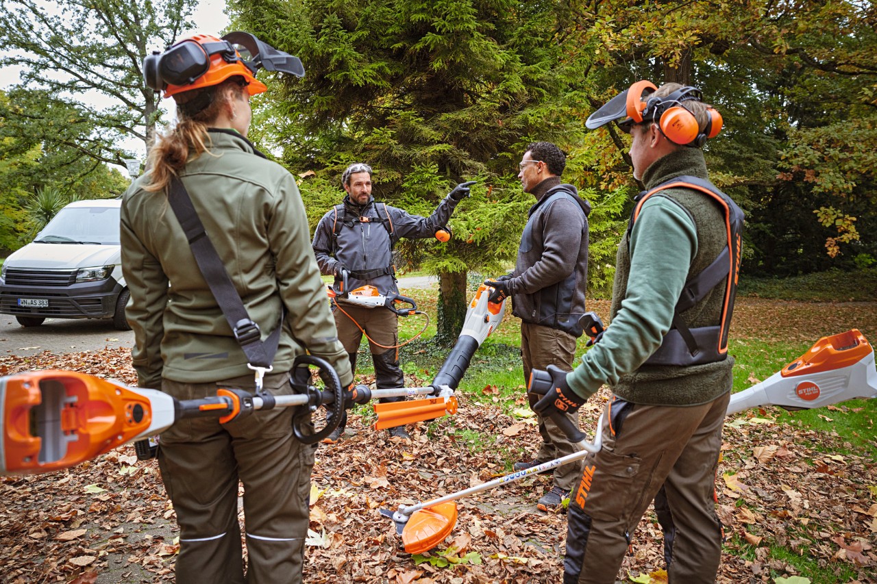 A gardening and landscaping team with STIHL professional cordless tools stands on a green area with leaves in the park