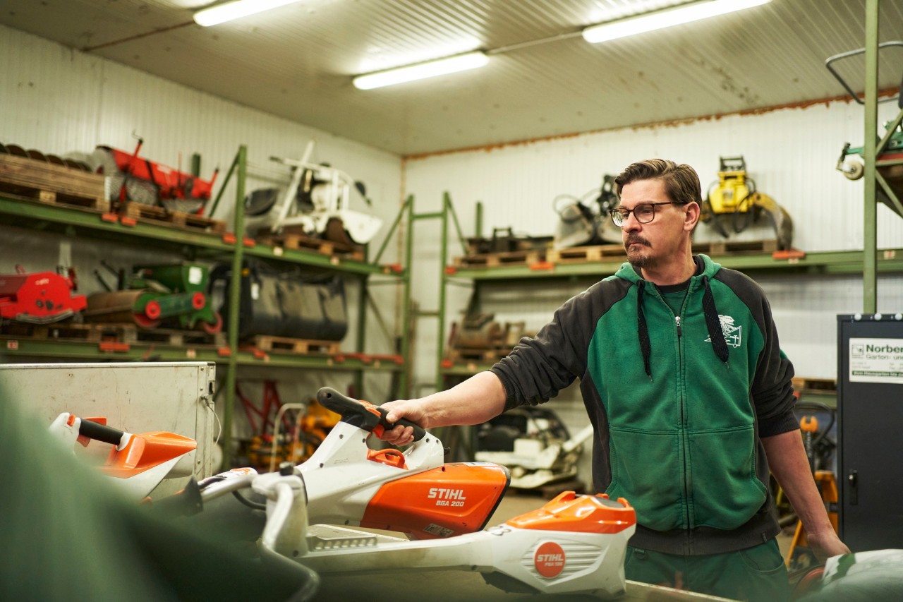 A man in a workshop stands behind a work surface containing STIHL battery power tools, and reaches for a STIHL BGA 200 battery leaf blower