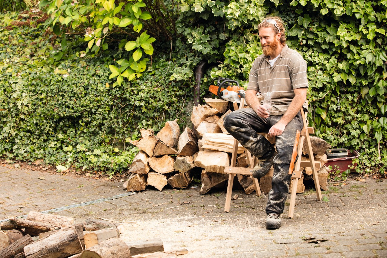 Man sitting on a sawhorse in front of a pile of split logs.
