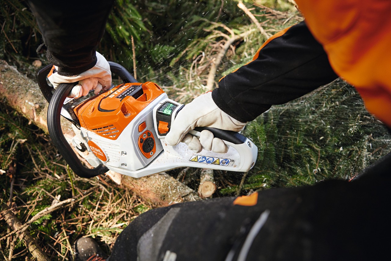 A STIHL professional under a roof structure saws a wooden beam with a battery chainsaw, while another worker uses a battery chainsaw in the background