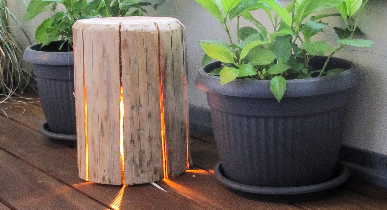 A DIY wooden lamp stands between two plant pots, with a string of lights lit up inside it