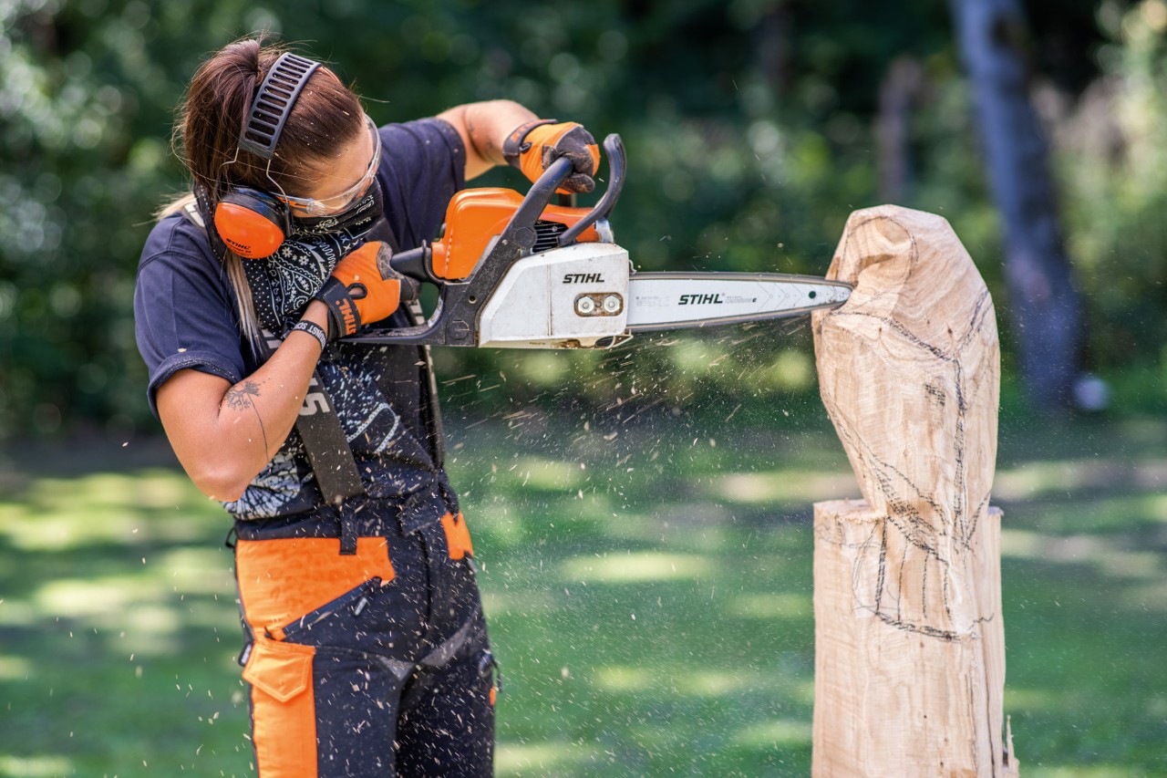 A woman wearing STIHL ear protection carving a wooden owl chainsaw