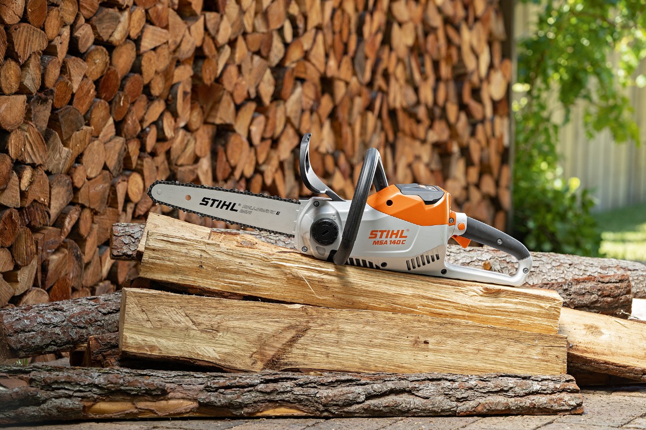 STIHL MSA 140 C-B cordless chainsaw on a pile of long logs, with neatly stacked firewood in the background