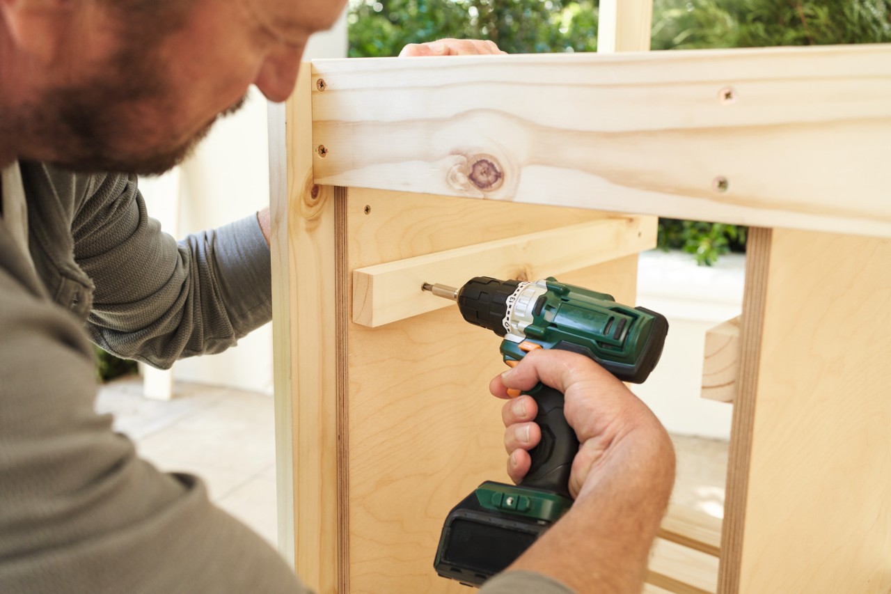 A man using screws to fix rails for a waste basket onto a potting bench
