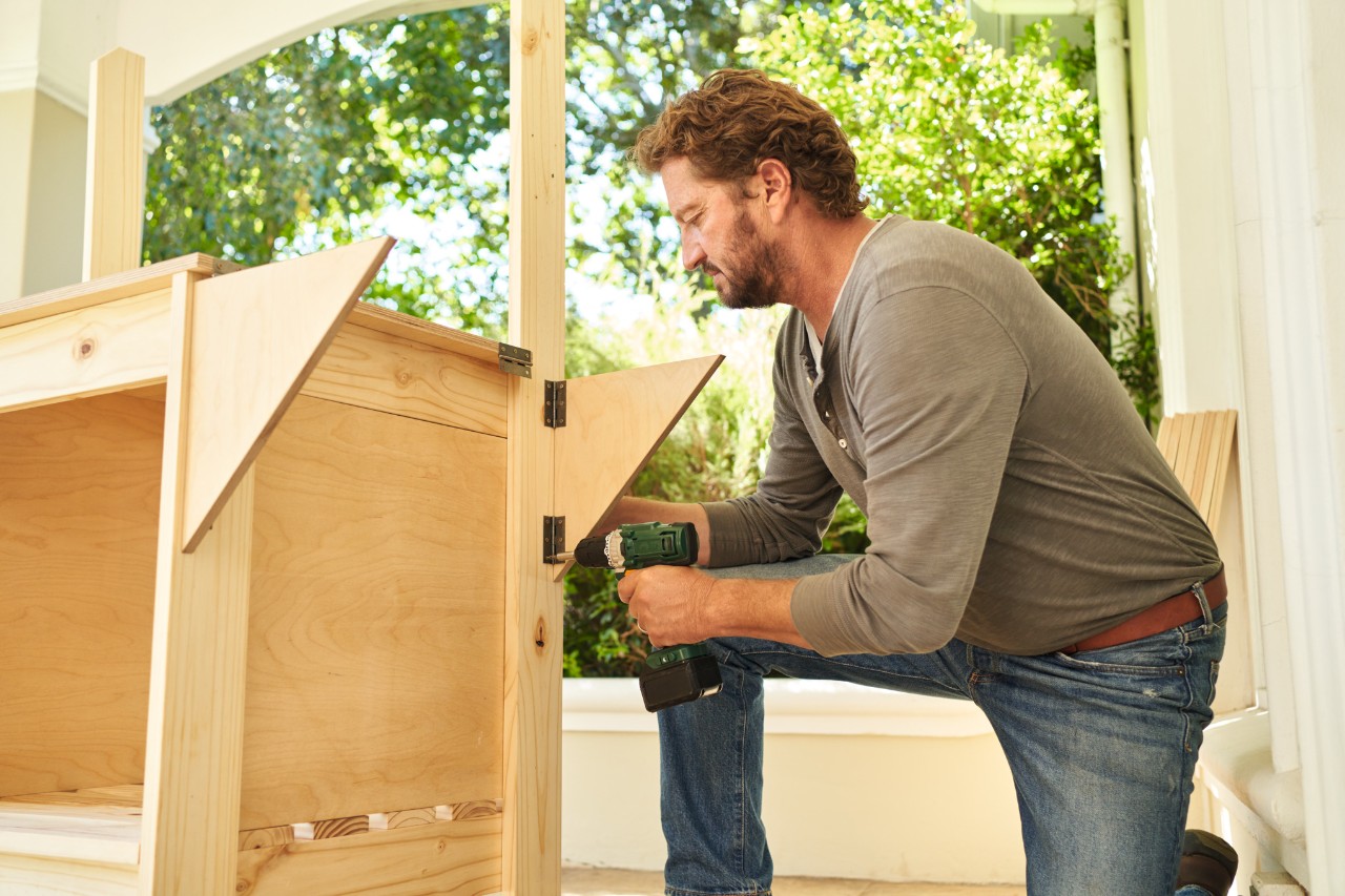 A man on a terrace using a screwdriver to attach hinges with bracket pieces to a planting table