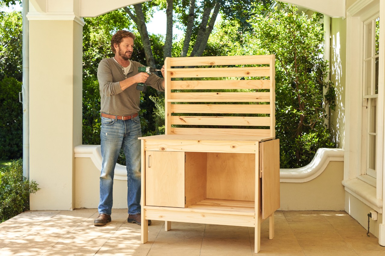 A man screwing shelf boards onto the top back panel of a potting bench 