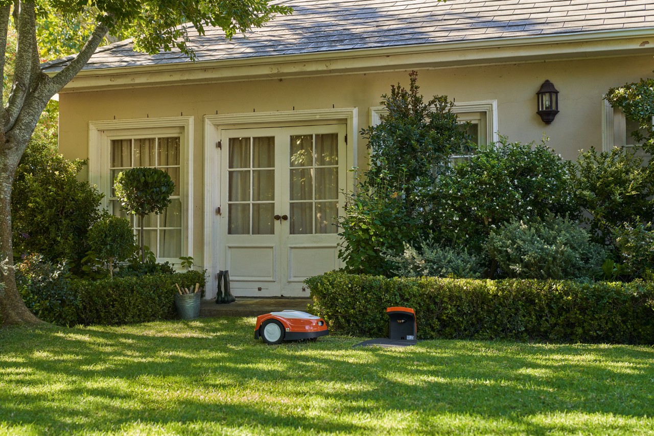 STIHL iMOW® robot mower next to the docking station on lush grass with hedges and trees near the entrance to a house