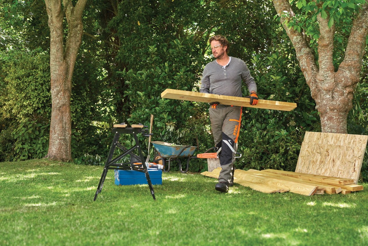 A person taking a plank of wood from a pile of materials under a tree