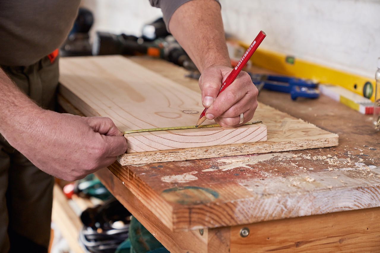 A person marking drill hole positions on a wooden board to build a DIY garden power tool rack