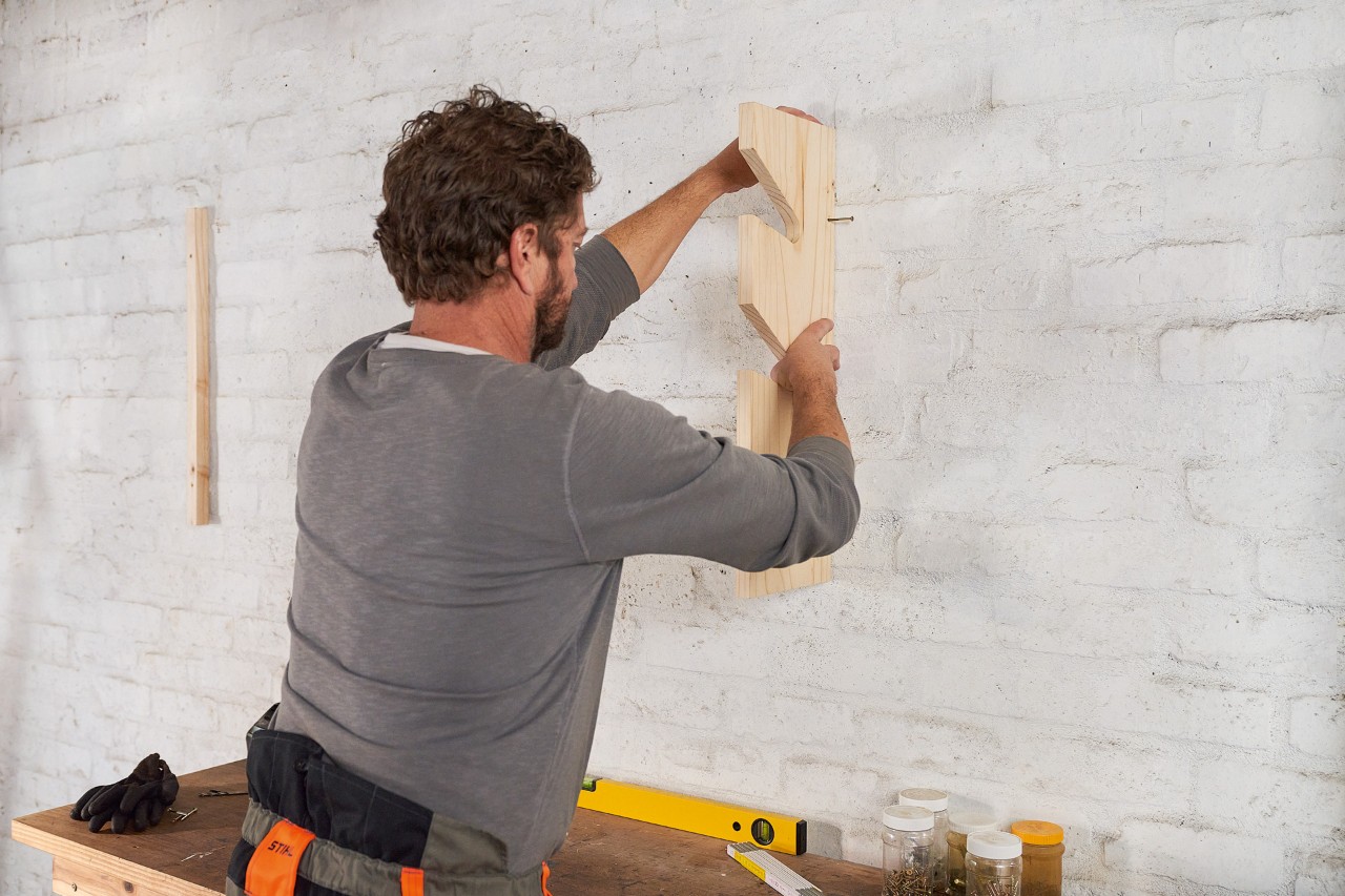 A man mounting a wooden batten to the wall with dowel pins to build a DIY garden power tool storage rack