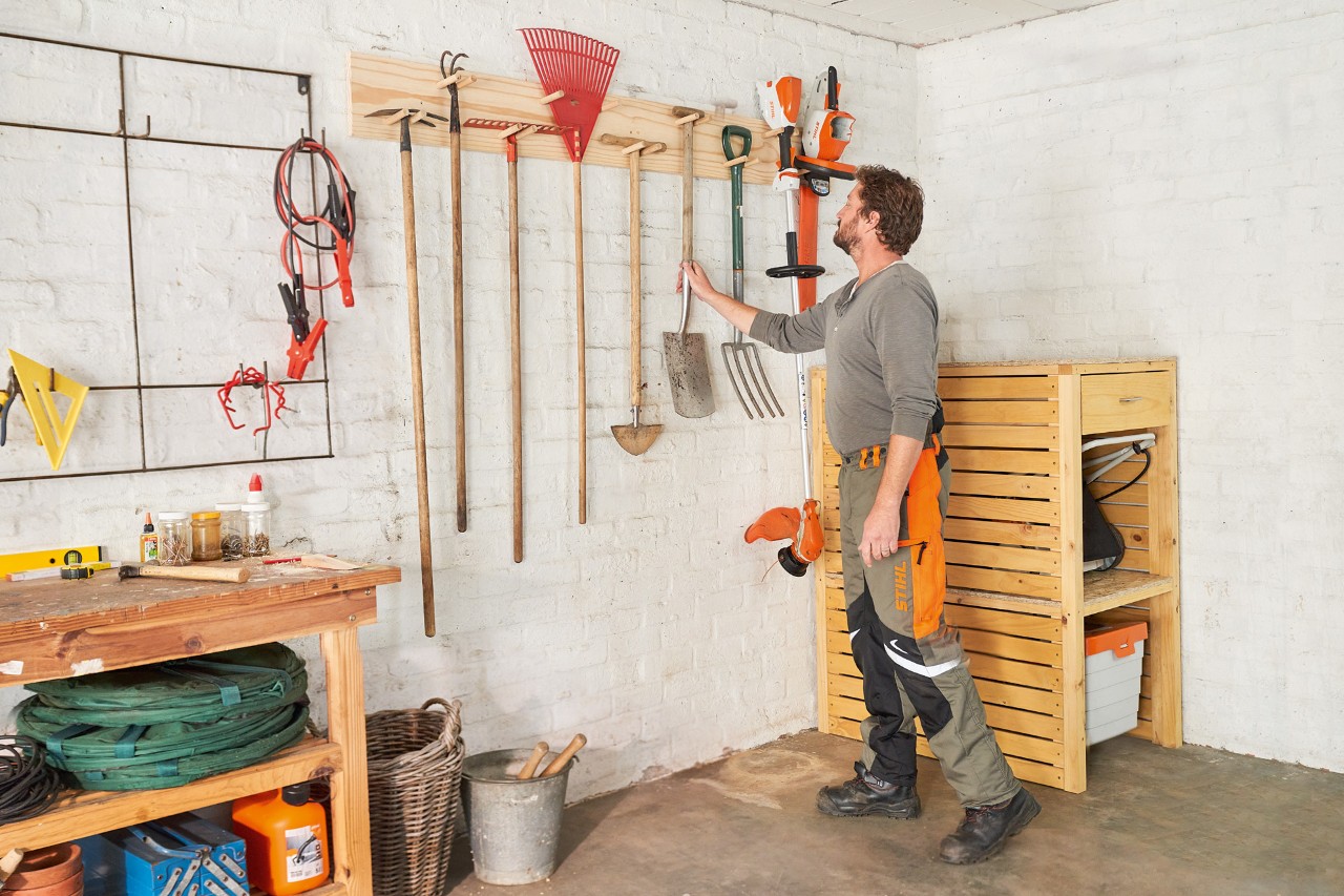 Man in STIHL work trousers using the DIY hanging garden tool rack to hang a shovel next to a STIHL grass trimmer