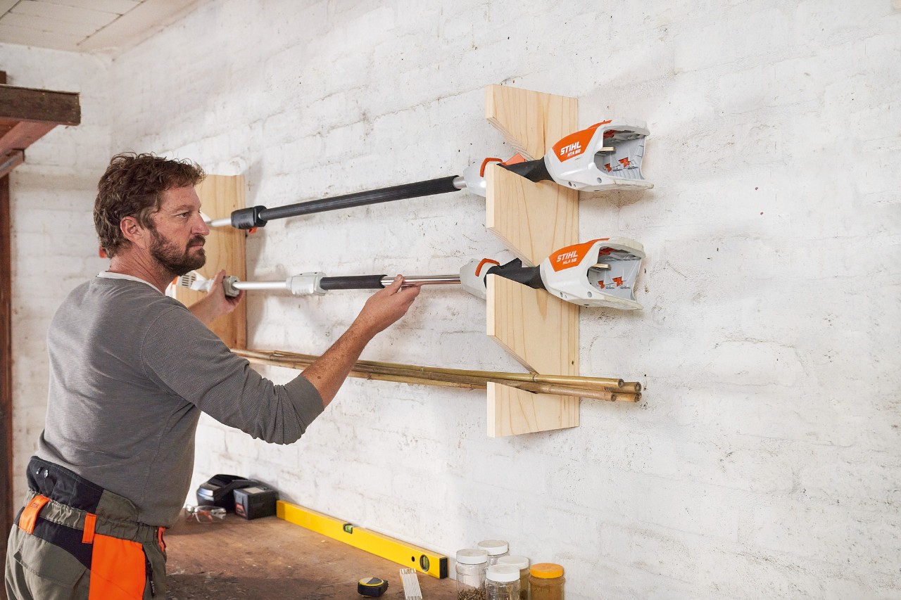 A man putting a STIHL brushcutter into a DIY garden power tool rack which is mounted on the wall