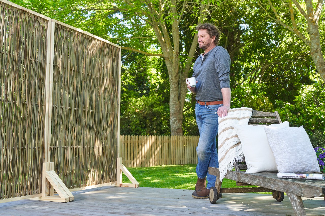 A man holding a coffee cup stands on the terrace, looking proudly at his DIY privacy screen