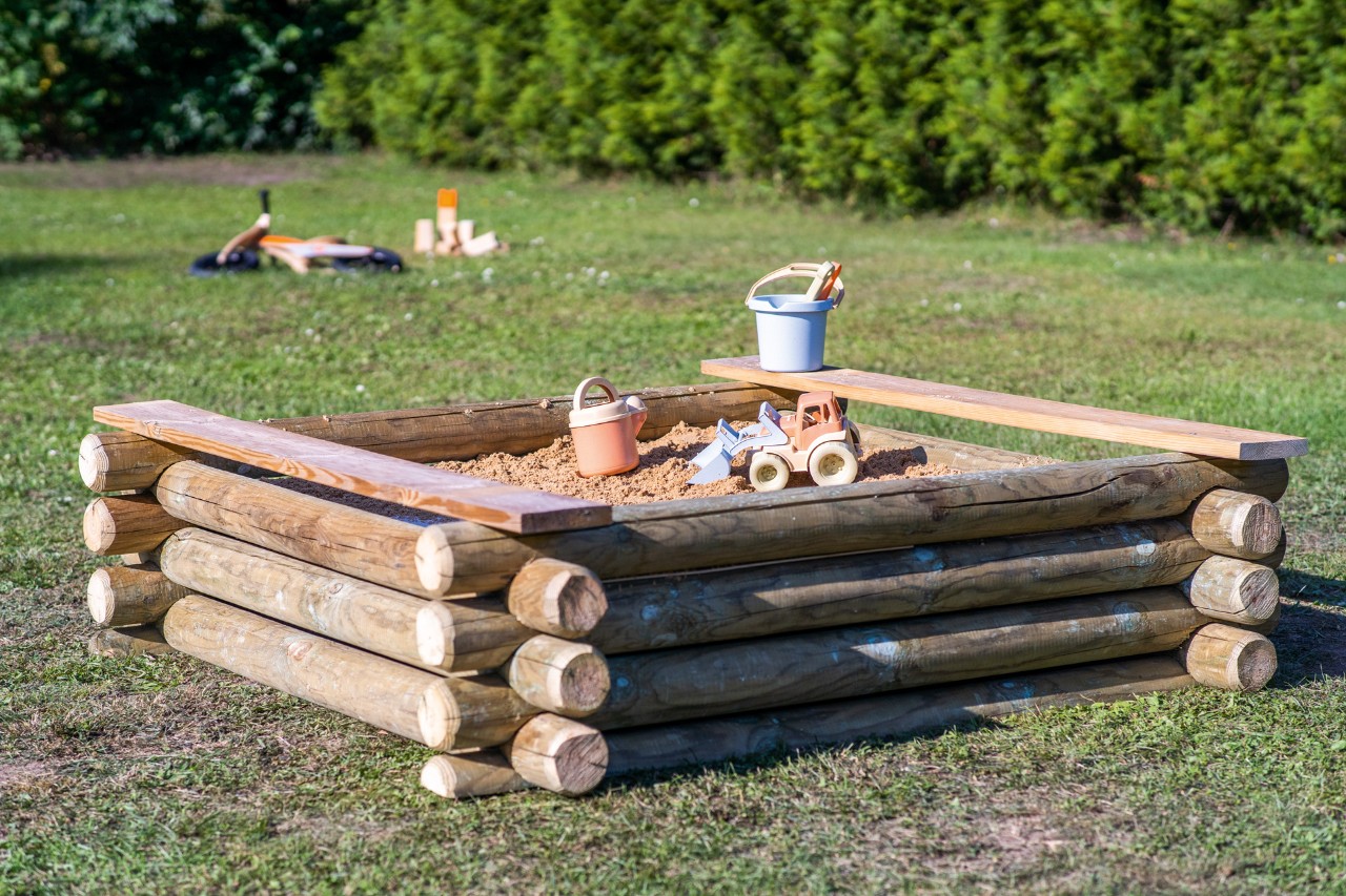 A DIY wooden sandpit with bench seats stands on a green lawn, with buckets and other toys inside it.
