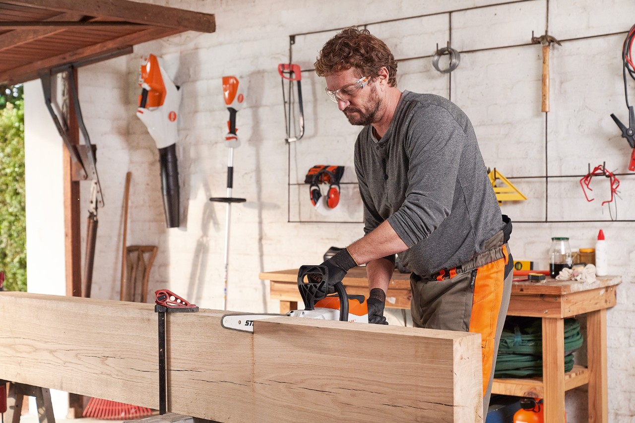 A man in a workshop wearing protective equipment and cutting a wooden plank with the STIHL cordless chainsaw MSA 140 C.