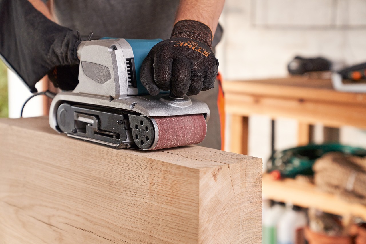 Close-up of a person wearing STIHL protective gloves while sanding wood in a workshop using a sander.