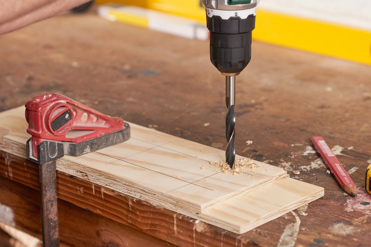 A cordless screwdriver being used to drill a hole into a thin wooden board to make a drilling template