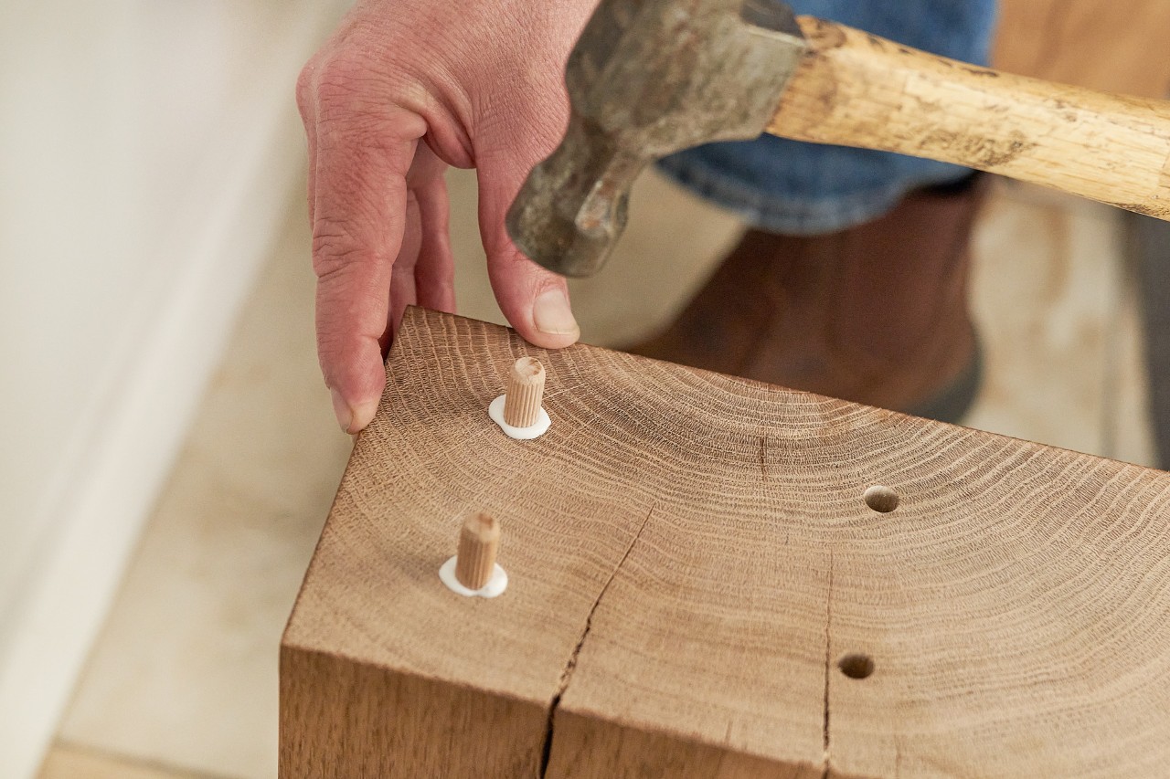 A person using a hammer to drive dowels into a wooden plank
