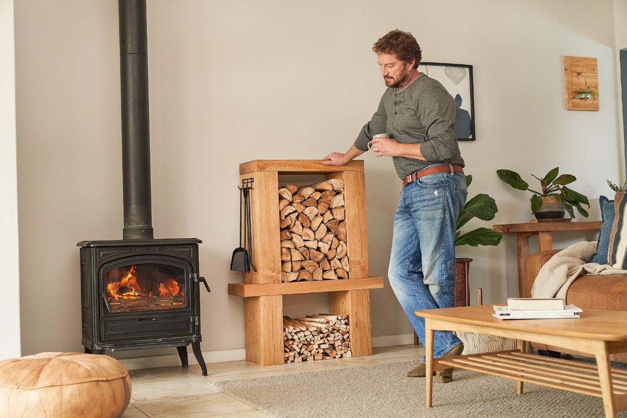 A man standing in a furnished room next to a DIY firewood rack with a cup in his hand