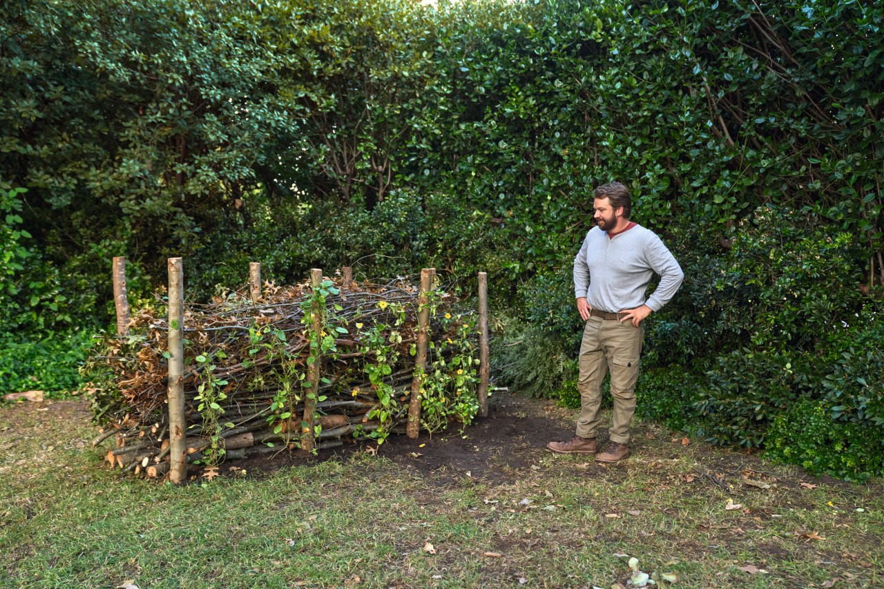 A man stands in front of a finished dead hedge with his arms folded and looks proudly at it