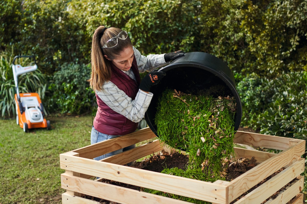 A woman empties the bucket filled with lawn clippings and shredded branches into the composter. RMA in the background.