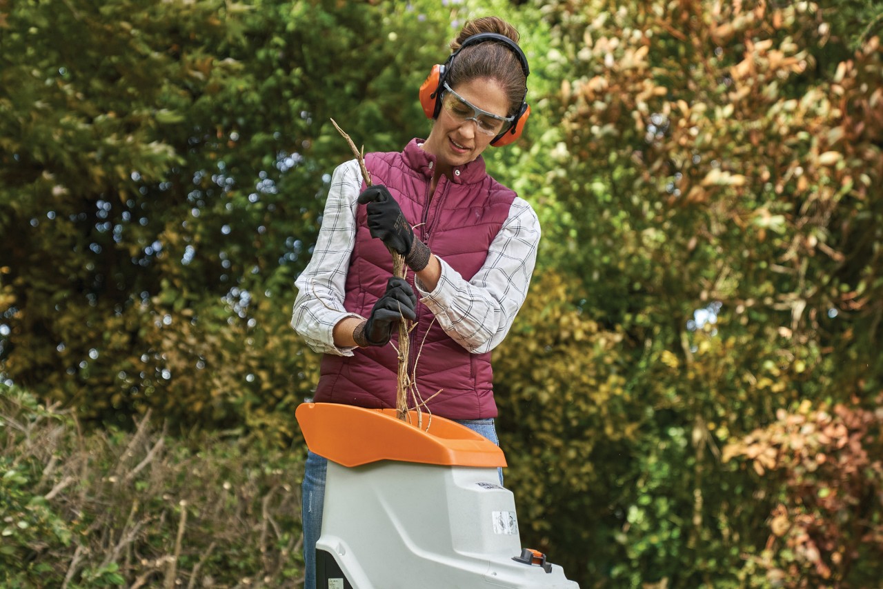 A woman wearing gloves, ear protection and glasses feeding branches into a garden shredder