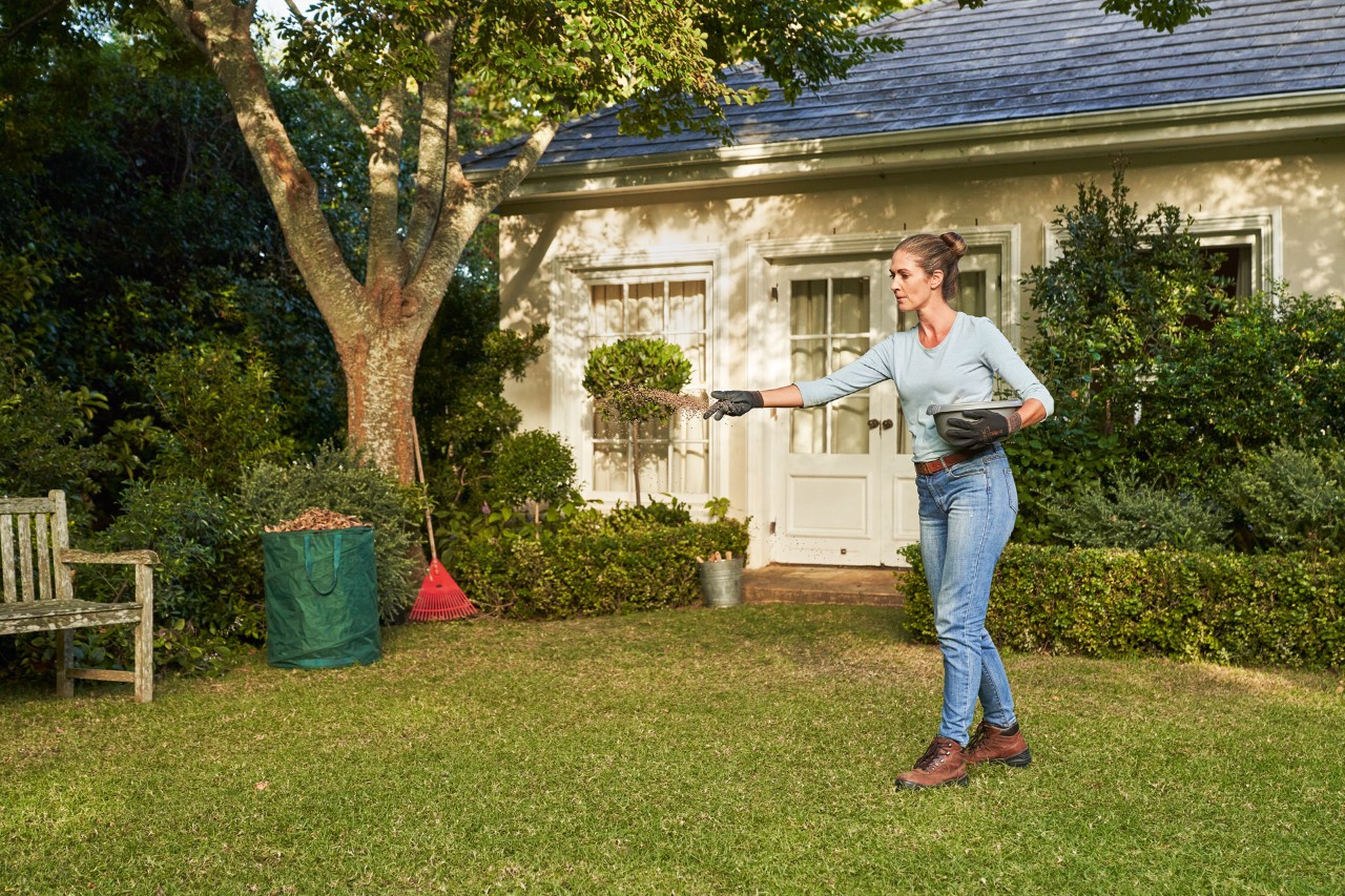 A woman wearing STIHL FUNCTION SENSOTOUCH gloves adds garden lime to the lawn, with bushes and a house in the background