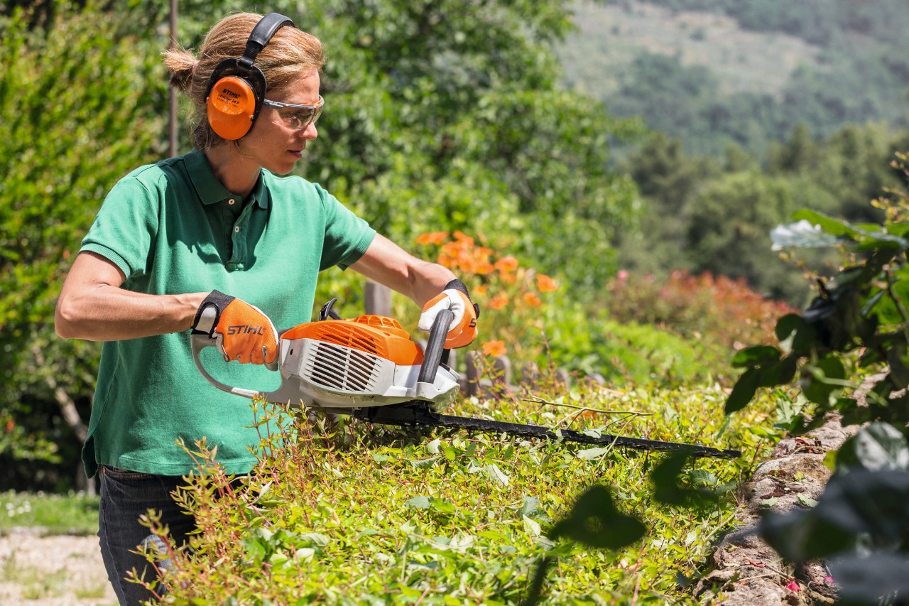 Woman wearing ear protection and safety glasses trimming a hedge with a STIHL HS 46 petrol hedge trimmer
