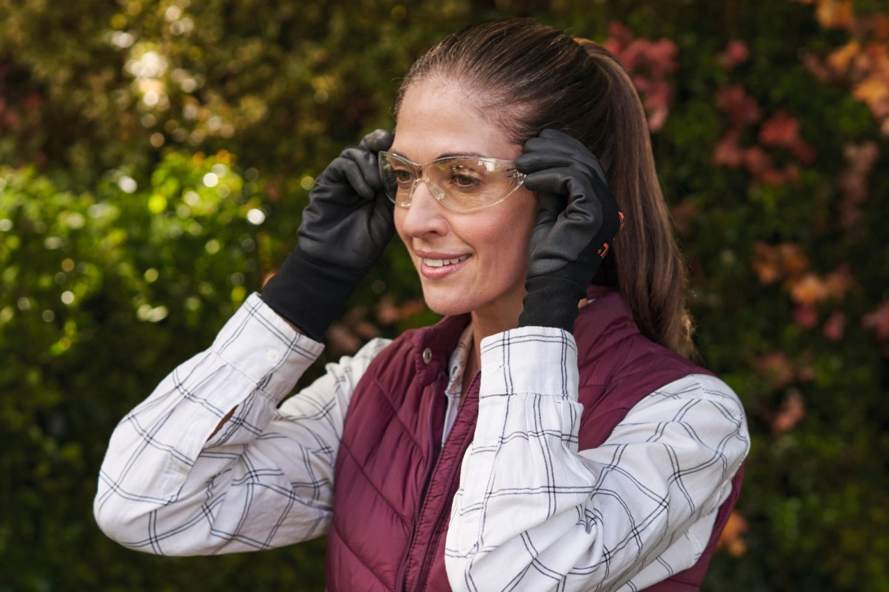 A smiling, brown-haired woman wearing protective gloves puts on safety glasses before pruning a thuja hedge