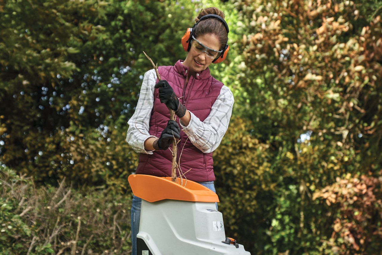 A woman in personal protective equipment shredding arborvitae hedge trimmings with trees in the background