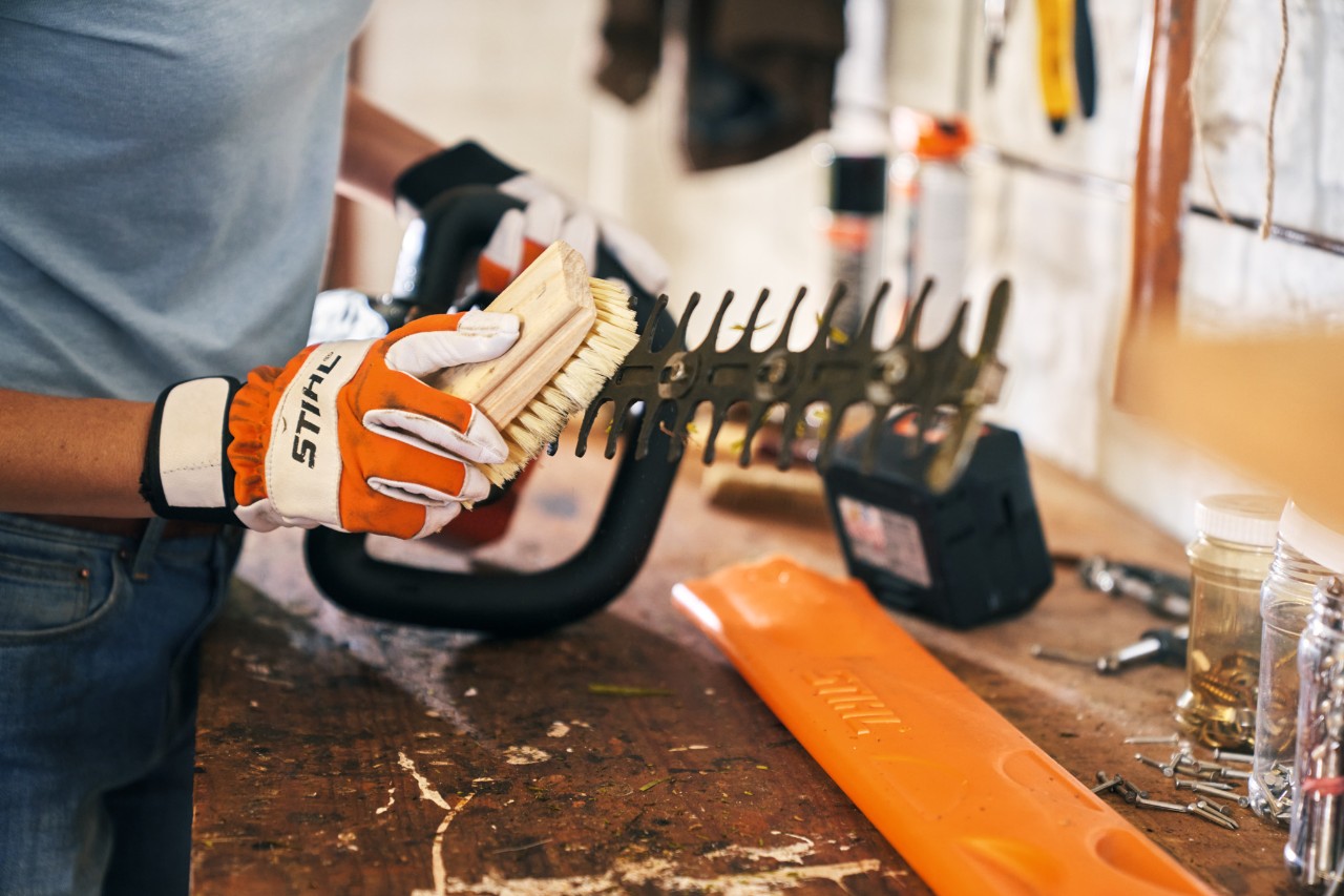 A person wearing protective gloves cleaning a hedge trimmer with a brush in a workshop 
