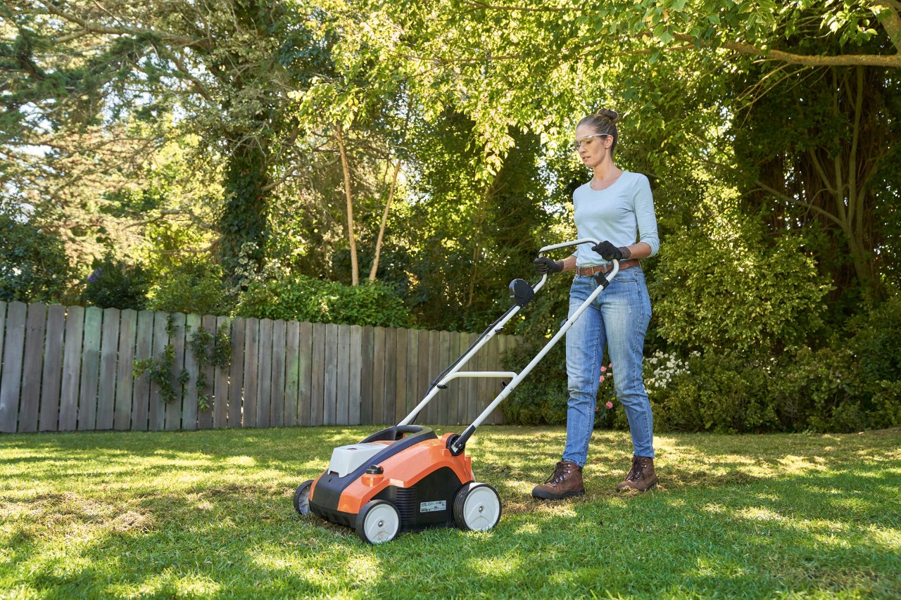 A woman wearing safety glasses and work gloves, aerating a garden lawn with a STIHL RLA 240 cordless lawn scarifier.