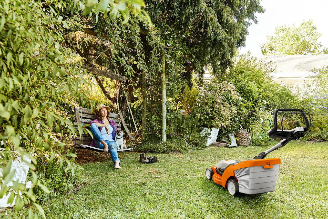 A woman relaxing on a garden swing next to a STIHL RMA 339 cordless lawn mower on a green plot