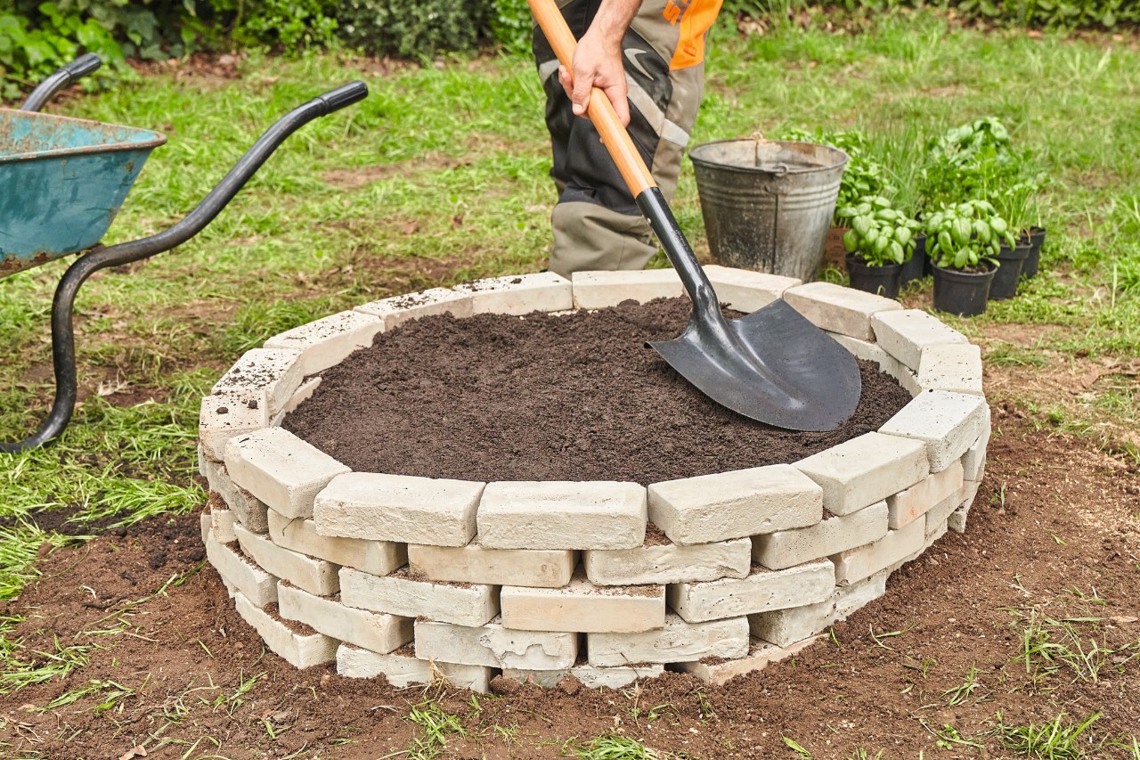 A person using a spade to evenly spread potting soil over the tree stump ringed by stones