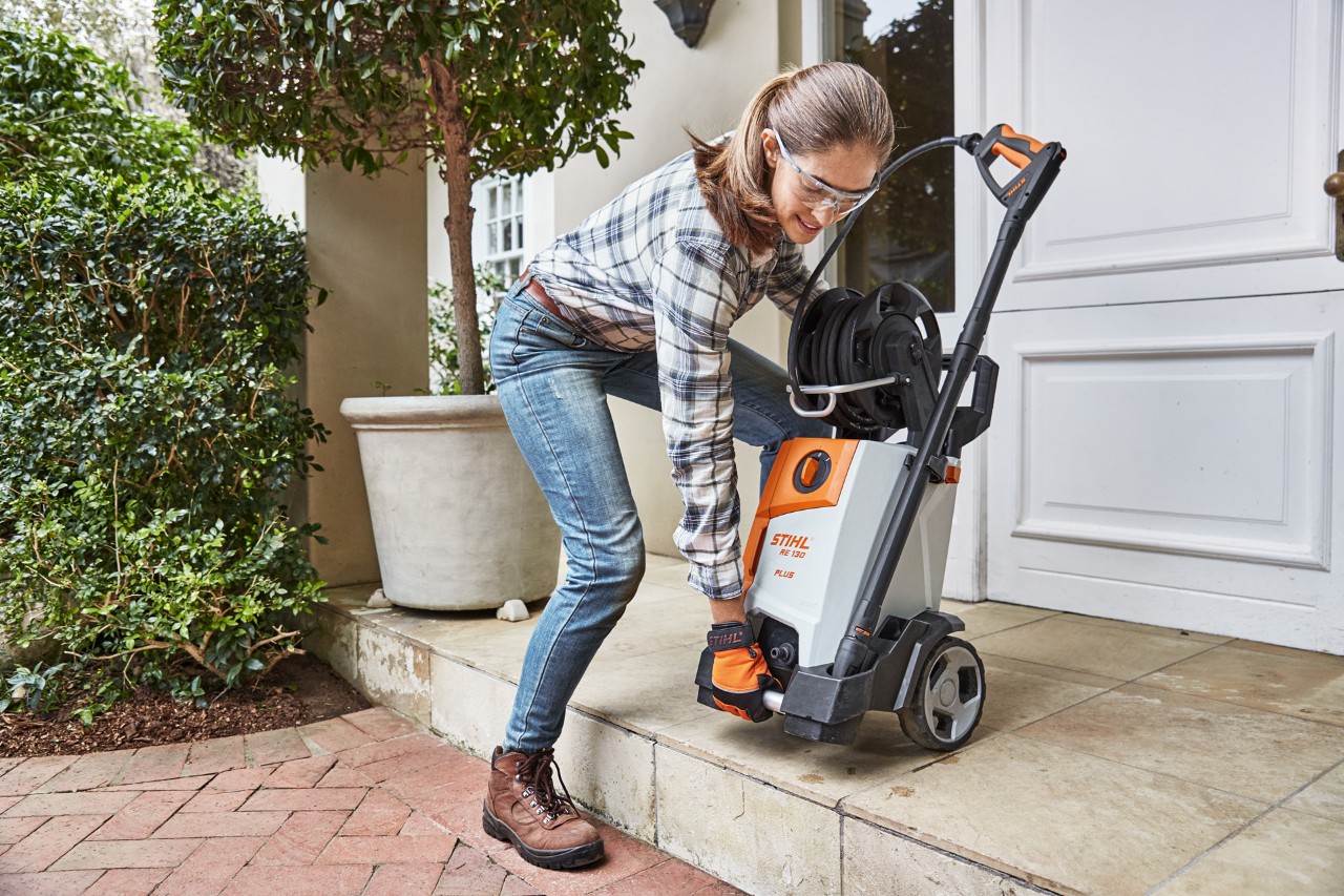 A woman lifting a STIHL RE 130 Plus high-pressure cleaner down a step to a brick path