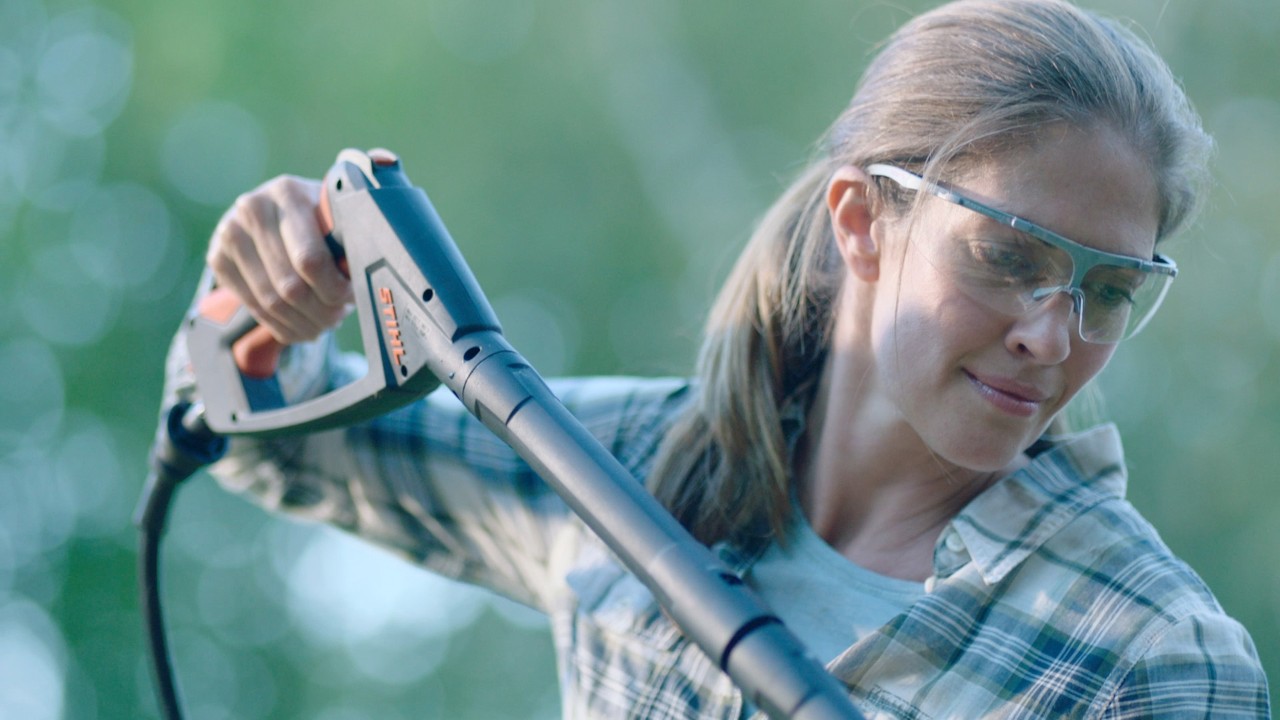 A woman wearing safety glasses holds up the handle of a STIHL RE 130 Plus high-pressure cleaner