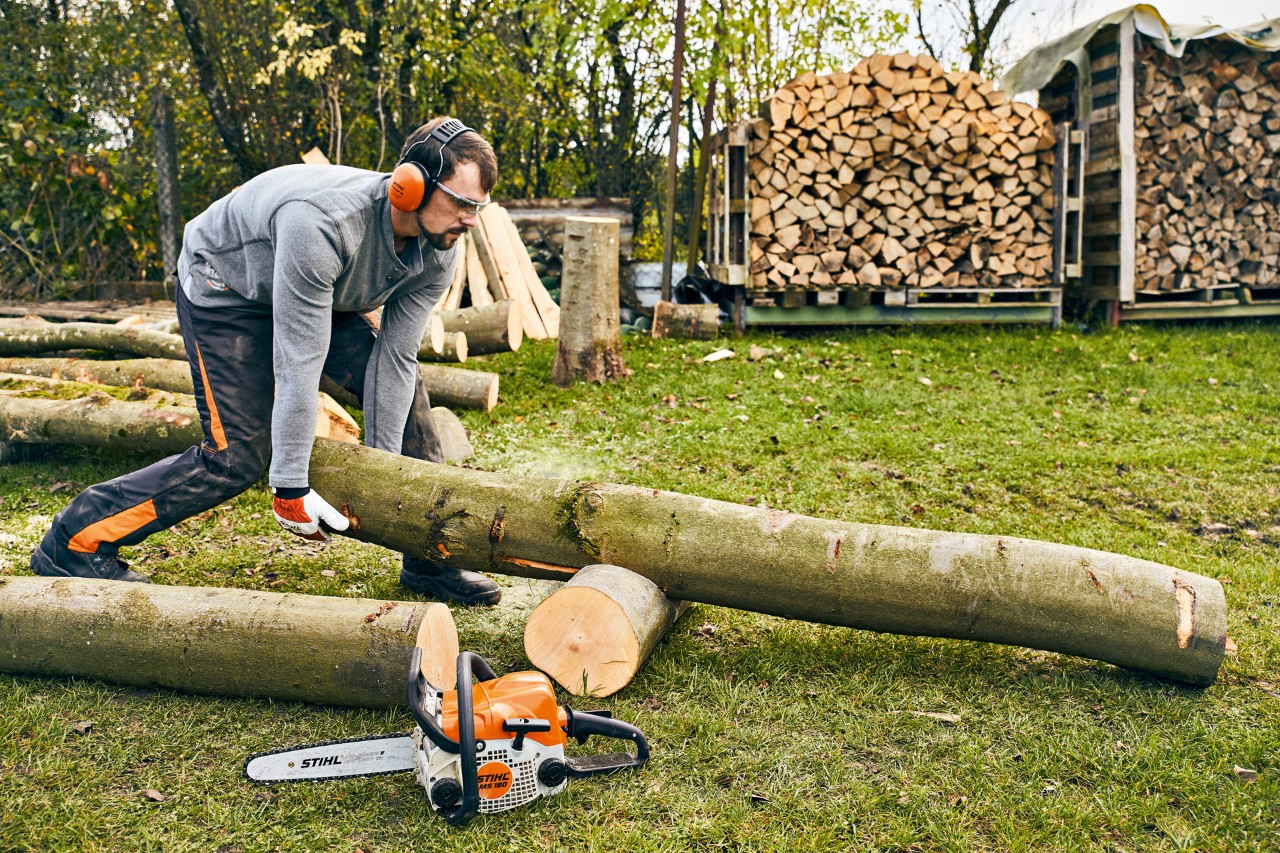 A tree trunk is repositioned on a cutting stand, with a STIHL MS 180 petrol-driven chainsaw on the ground in front.