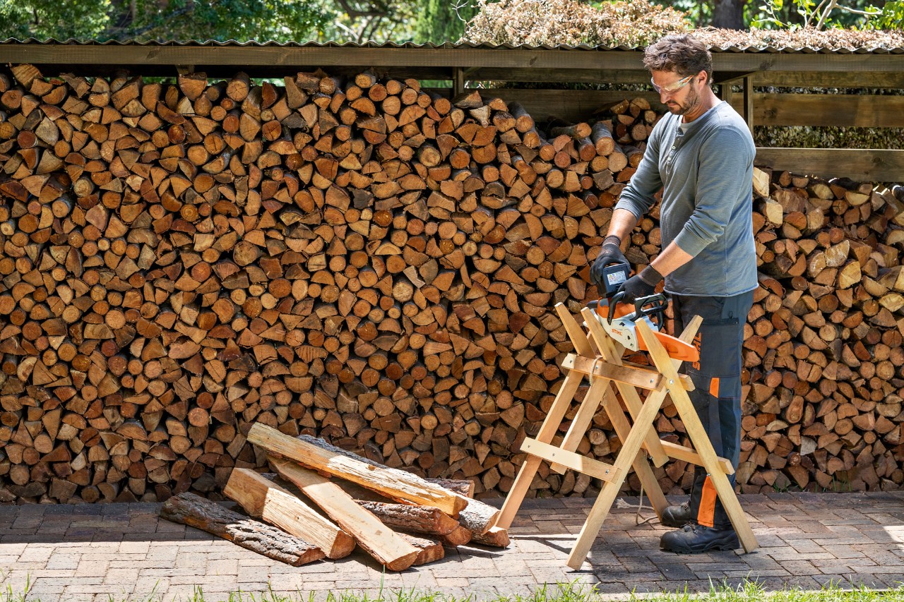 A man wearing chainsaw PPE is lifting a STIHL chainsaw off a saw horse beside a large stack of firewood.