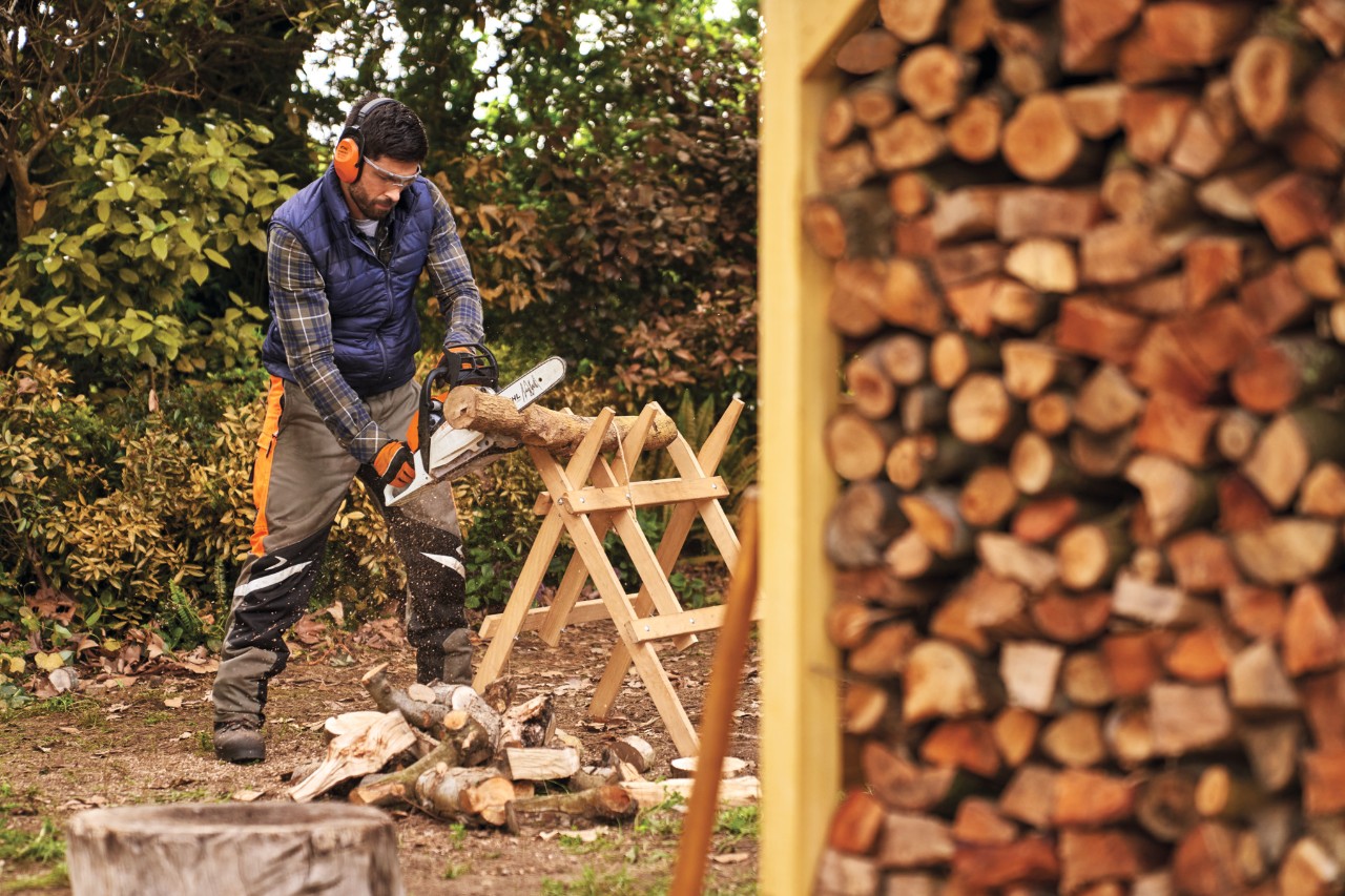 Man wearing protective equipment sawing logs for the outdoor log storage shed