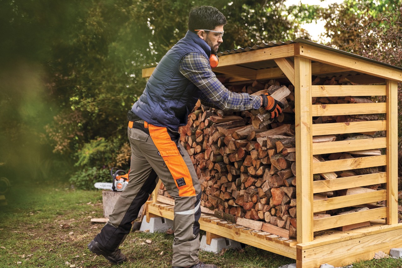 A man in protective clothing stacking firewood in a DIY log storage shed