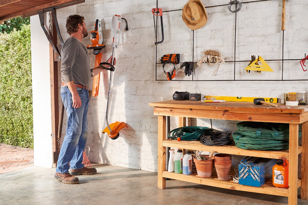 A man in a garage reaching for a STIHL electric brushcutter on the wall, with a STIHL cordless hedge trimmer beside it 