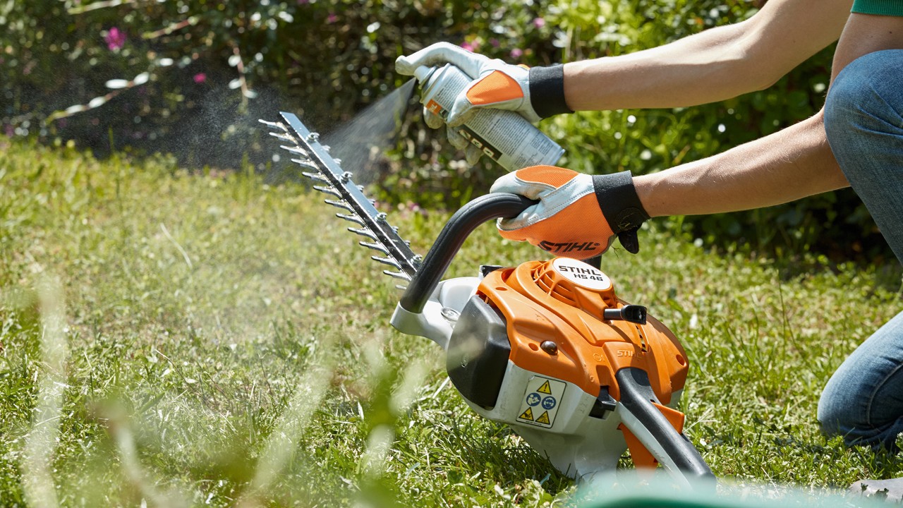A STIHL HSA 82 T petrol hedge trimmer on a lawn with someone spraying resin solvent onto the cutting attachment