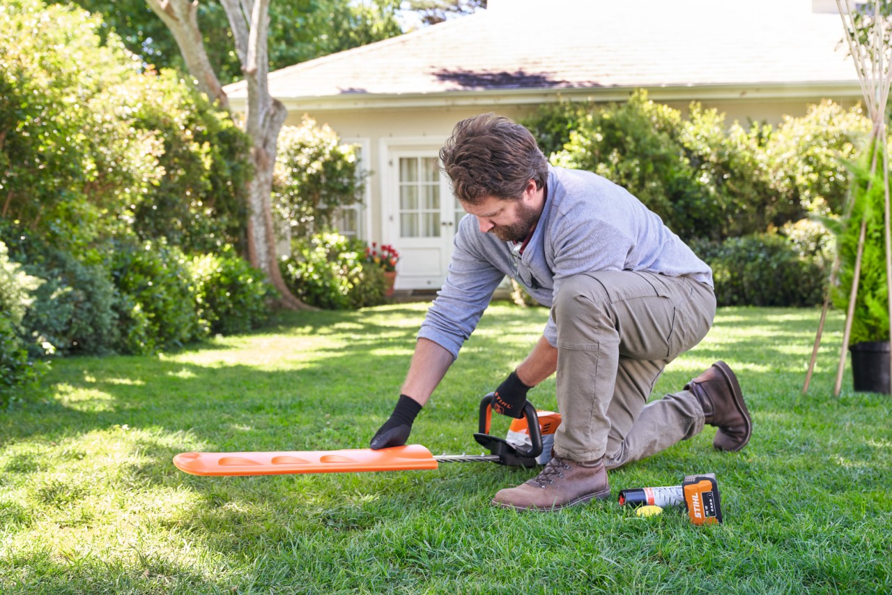 A man kneeling on the lawn removes the protective cover from a STIHL hedge trimmer 