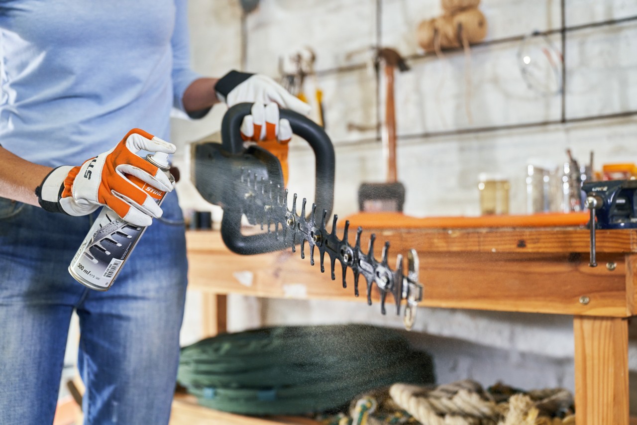 Woman wearing protective gloves sprays a STIHL HSA 56 battery hedge trimmer with cleaning spray for maintenance