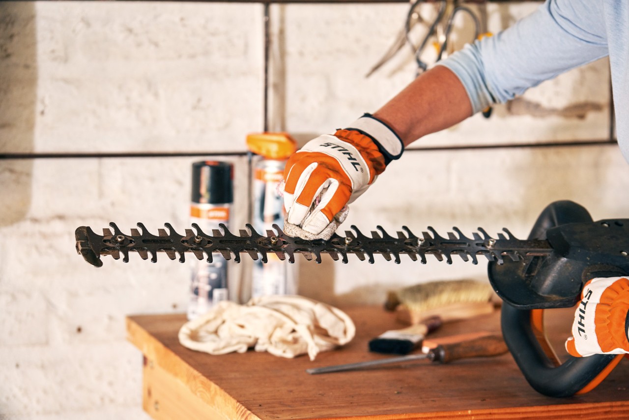A person wearing gloves holds a hedge trimmer on a workbench while using a cloth to clean the cutting apparatus