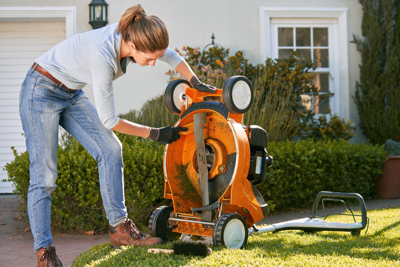 A woman cleaning the underside of a STIHL RM 448 T-C petrol lawn mower with a spatula