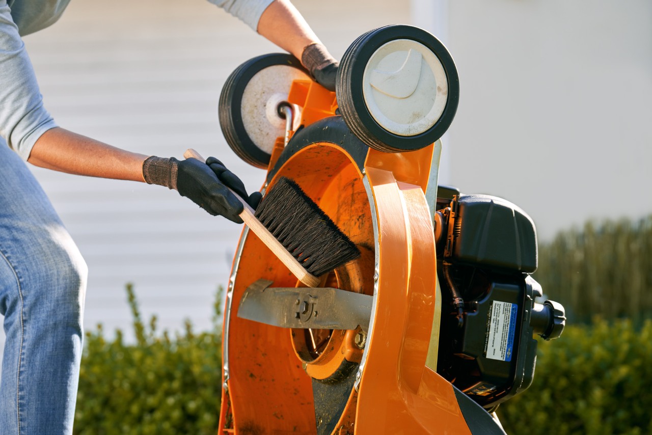 View of the underside of a lawn mower being held by a person wearing gloves and using a brush to clean near the blade