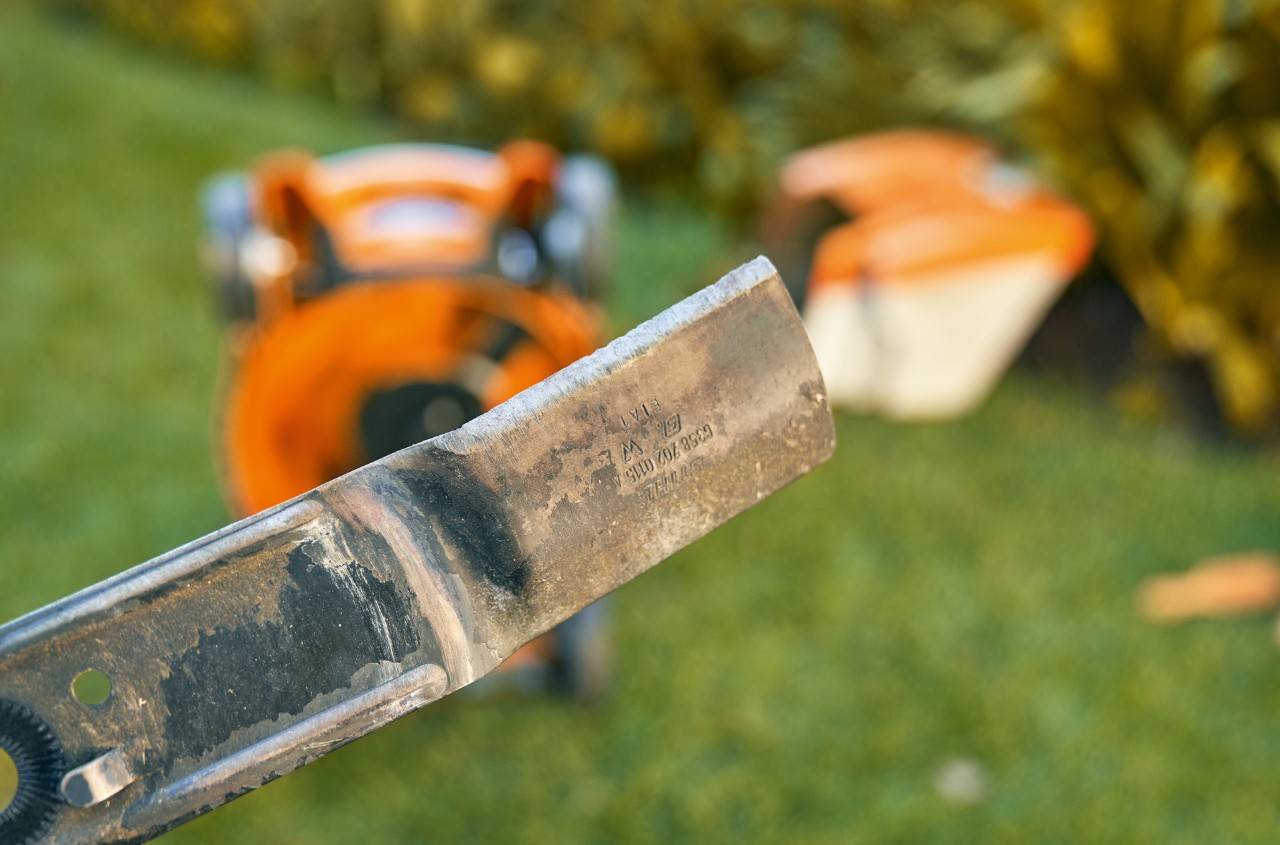 Close-up view of a cutting edge on a lawn mower blade, with an out-of-focus background of a lawn mower on grass